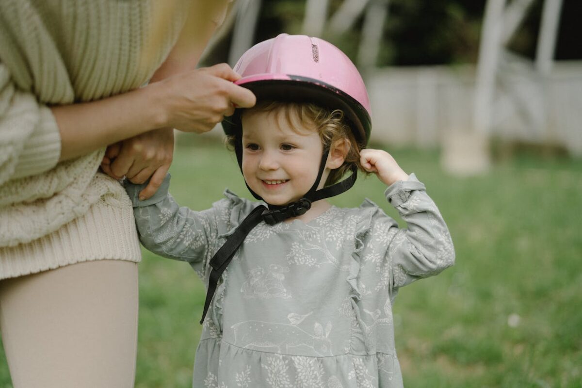Smiling young girl wearing a pink helmet, standing outdoors on grass.